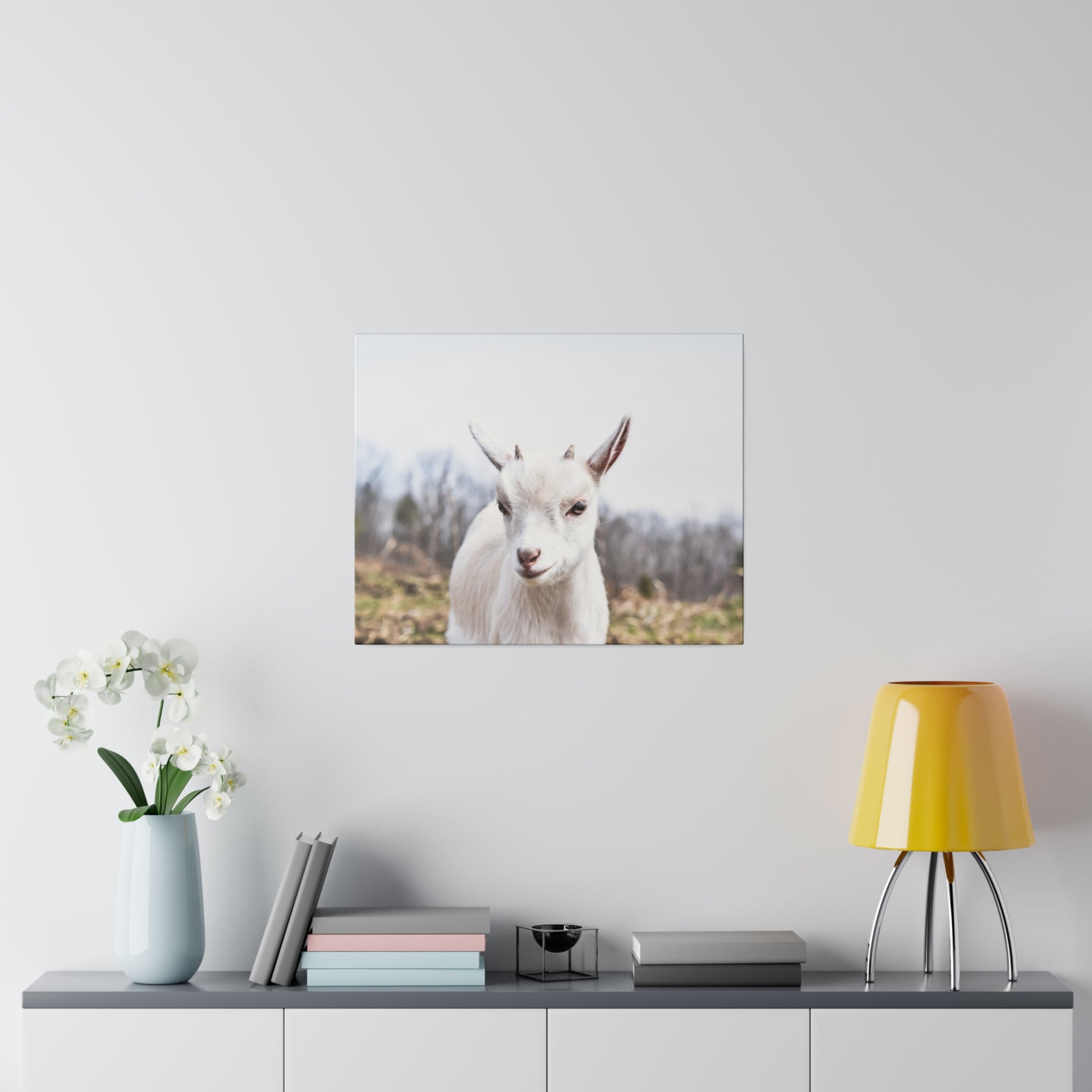 White goat portrait mounted on a light wall above a console table with a pale blue vase of orchids, stacked books, and a yellow lamp.