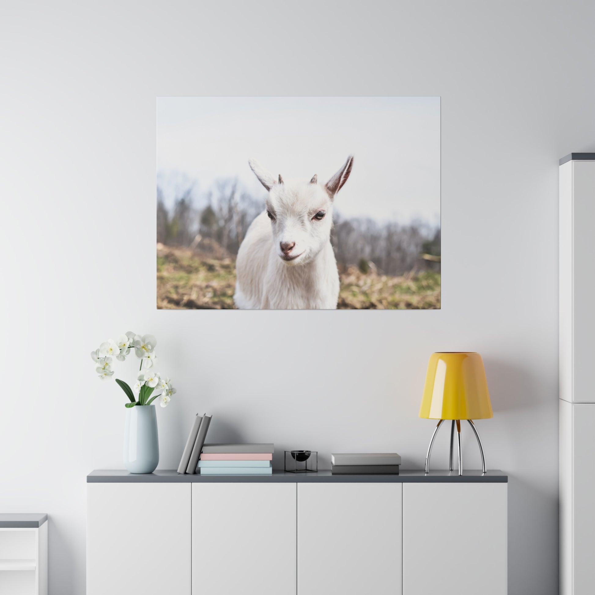White goat portrait mounted on a light gray wall above a cabinet with a blue vase, books, and a yellow table lamp.
