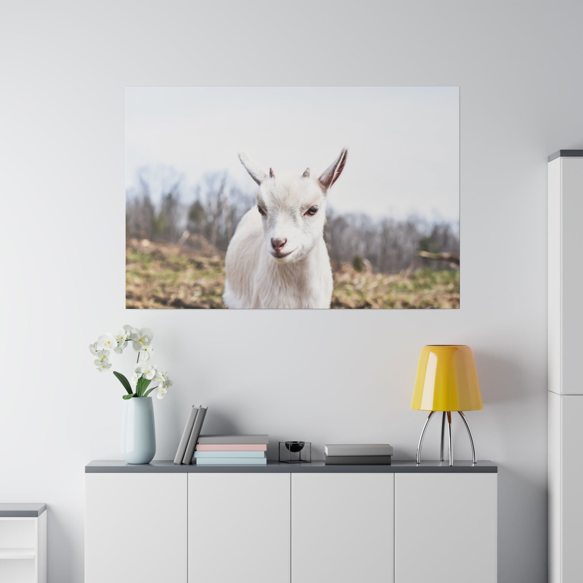 Large wall art of a white goat facing forward hangs above a gray cabinet with a yellow lamp and a vase of white orchids.