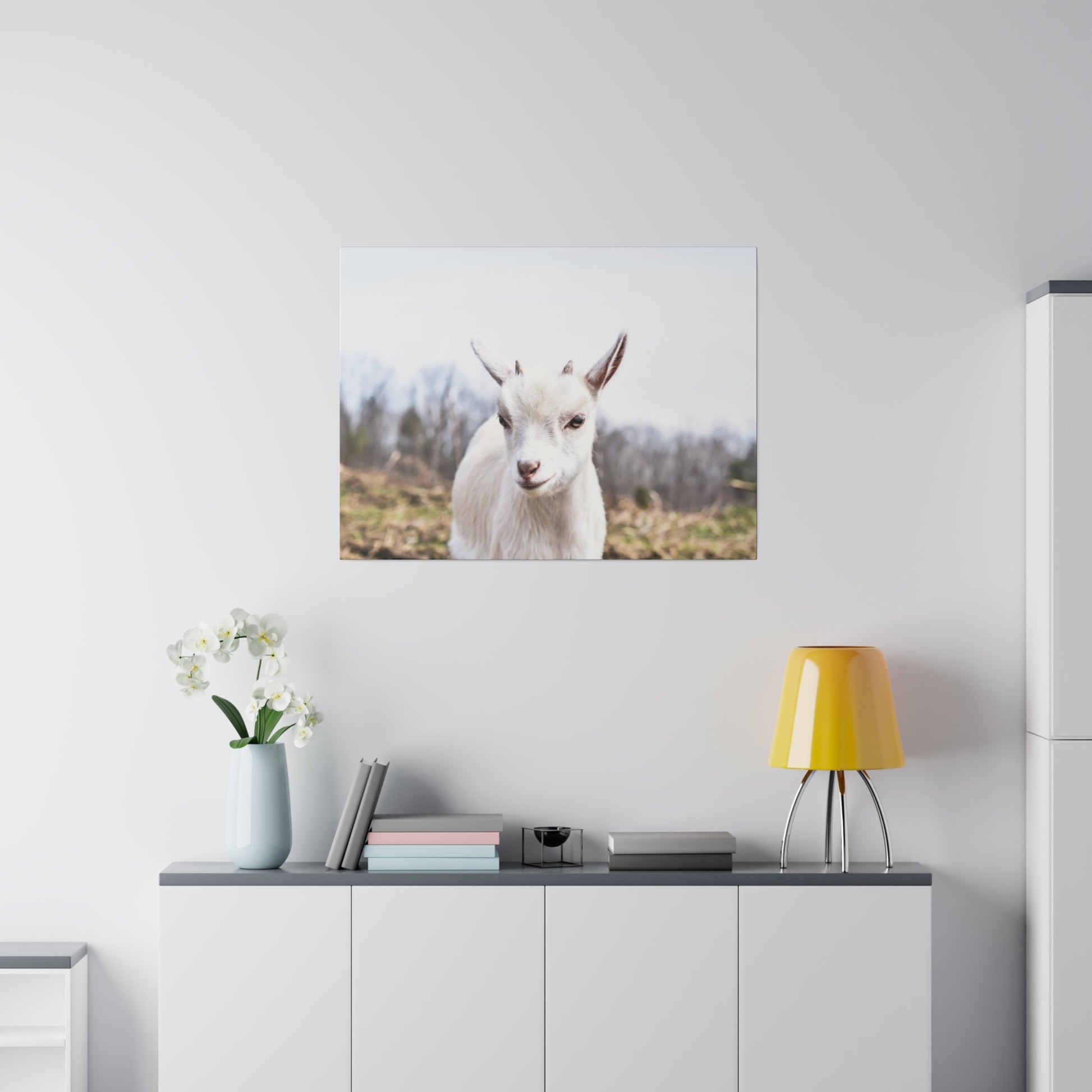Portrait of a white goat on a wall above a white cabinet, with a yellow lamp, books, and a vase of white orchids.
