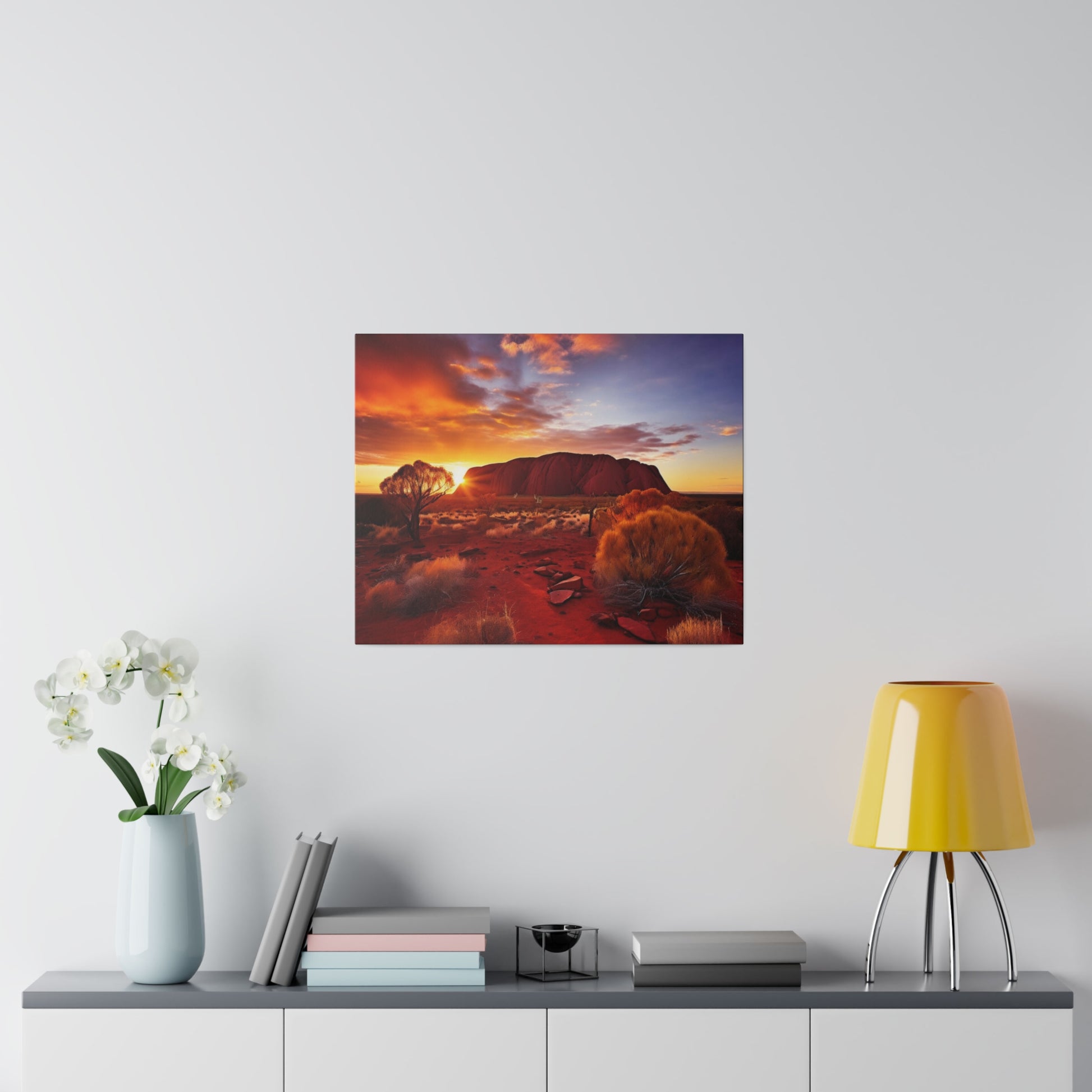 Square framed landscape photo of Uluru at sunset with orange rocks and sparse trees on a white wall.