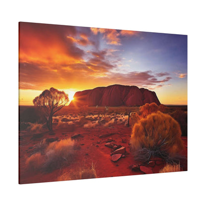 Desert landscape at sunset featuring Uluru rock formation, orange sky, and sparse red desert vegetation in the foreground.