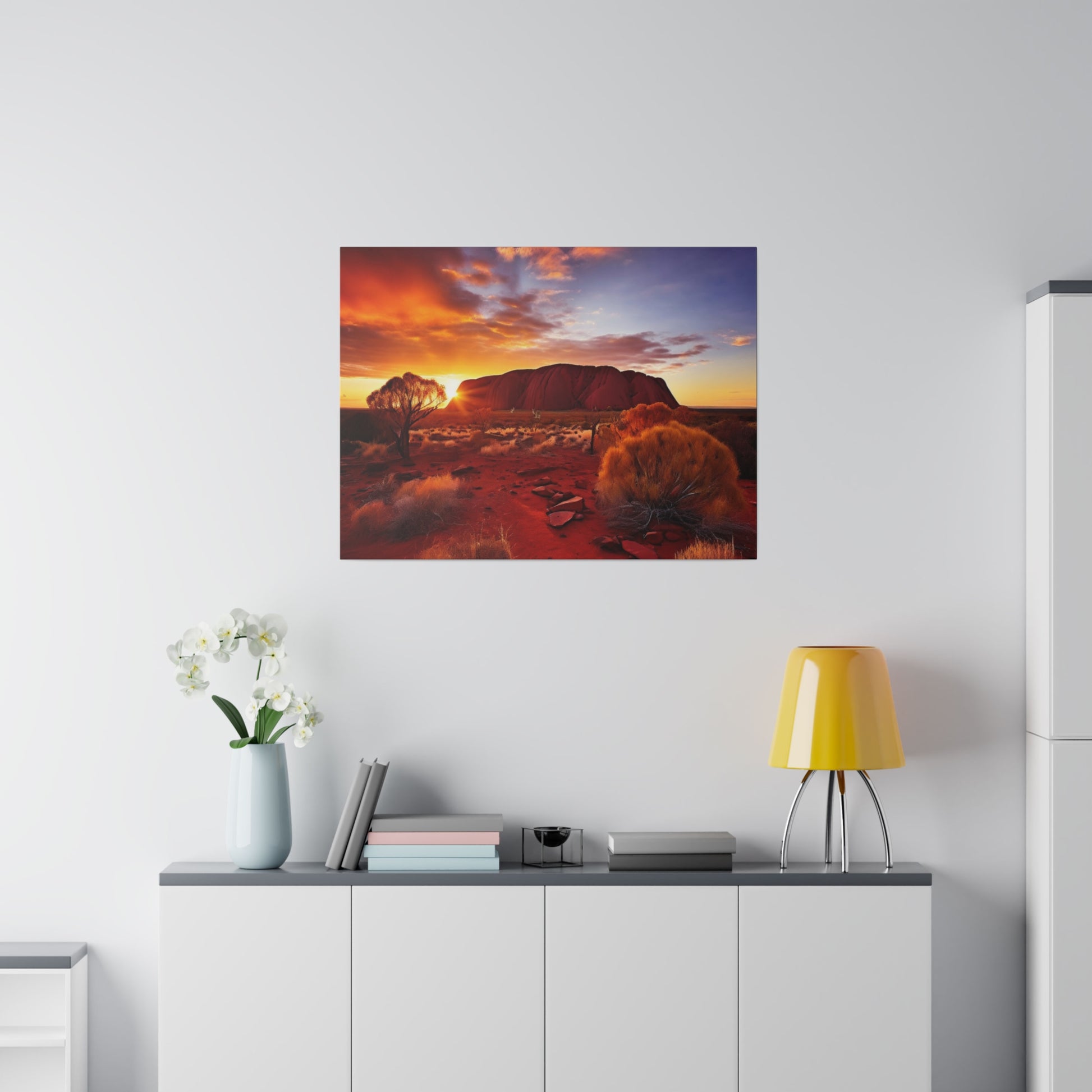 Desert landscape photo of Uluru at sunset hung on a white wall above a sideboard with a yellow lamp and vase.