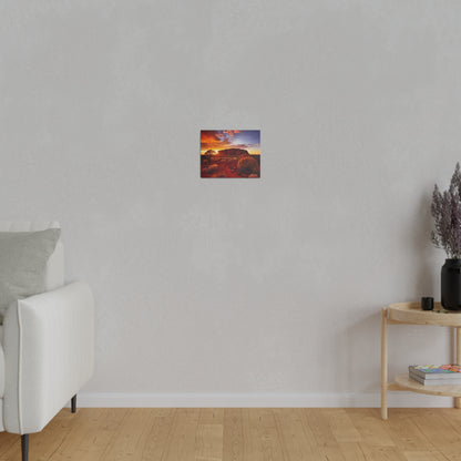 Small rectangular wall art depicting a desert landscape with an orange sunset, hung on a light gray wall above a wooden floor.