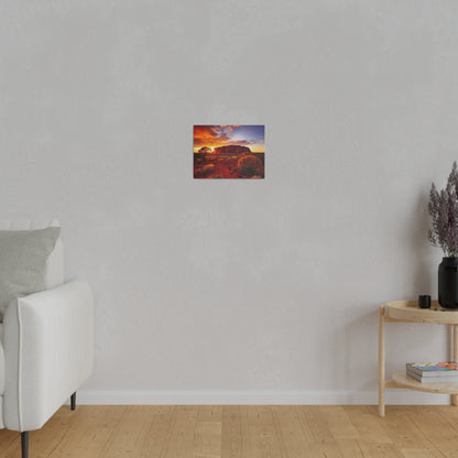 Small rectangular canvas showing a desert landscape with red rocks and a sunset, mounted on a light gray wall above a sofa in a minimalist