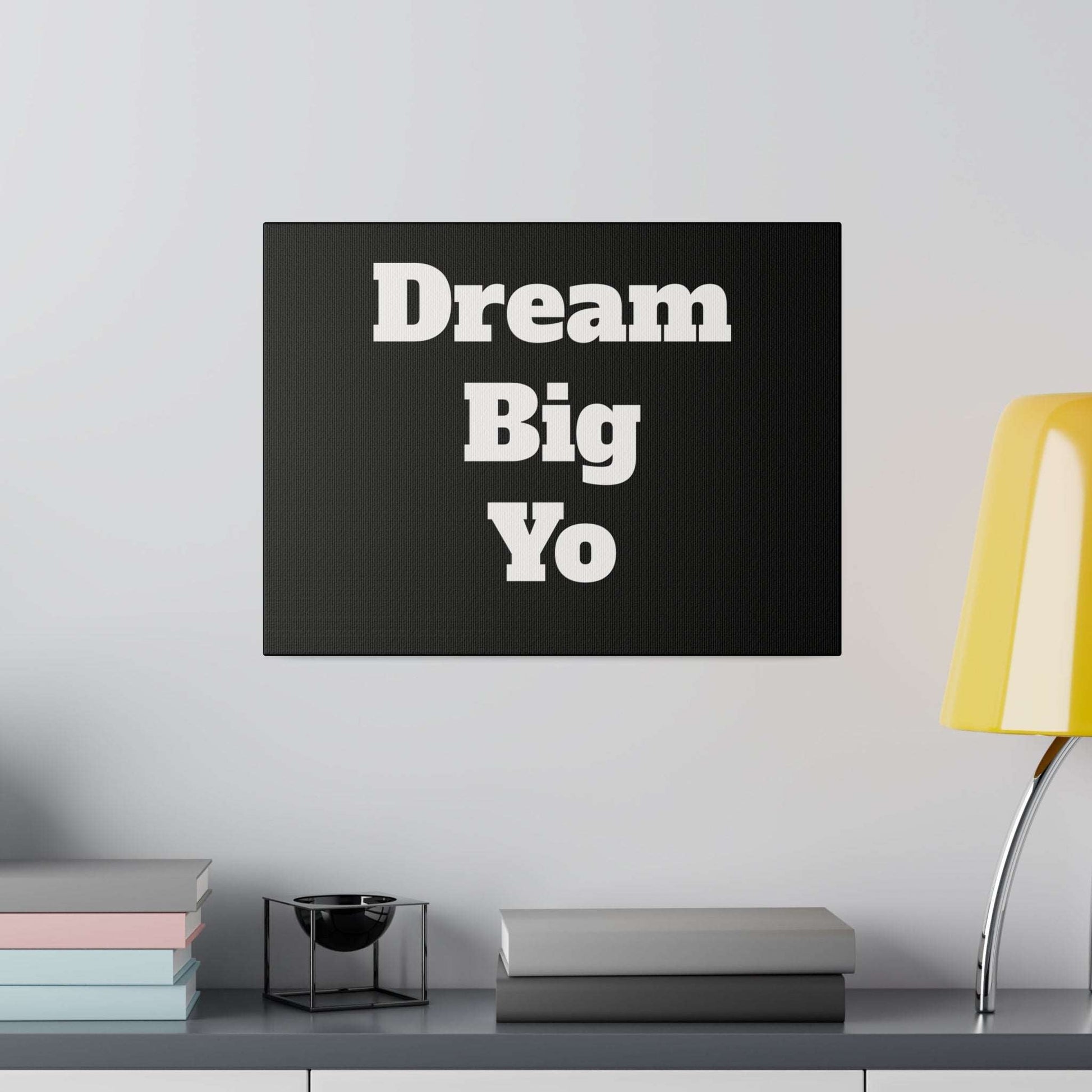 Black canvas wall art with bold white "Dream Big Yo" text, hung above a shelf with pastel books and a yellow curved lamp.