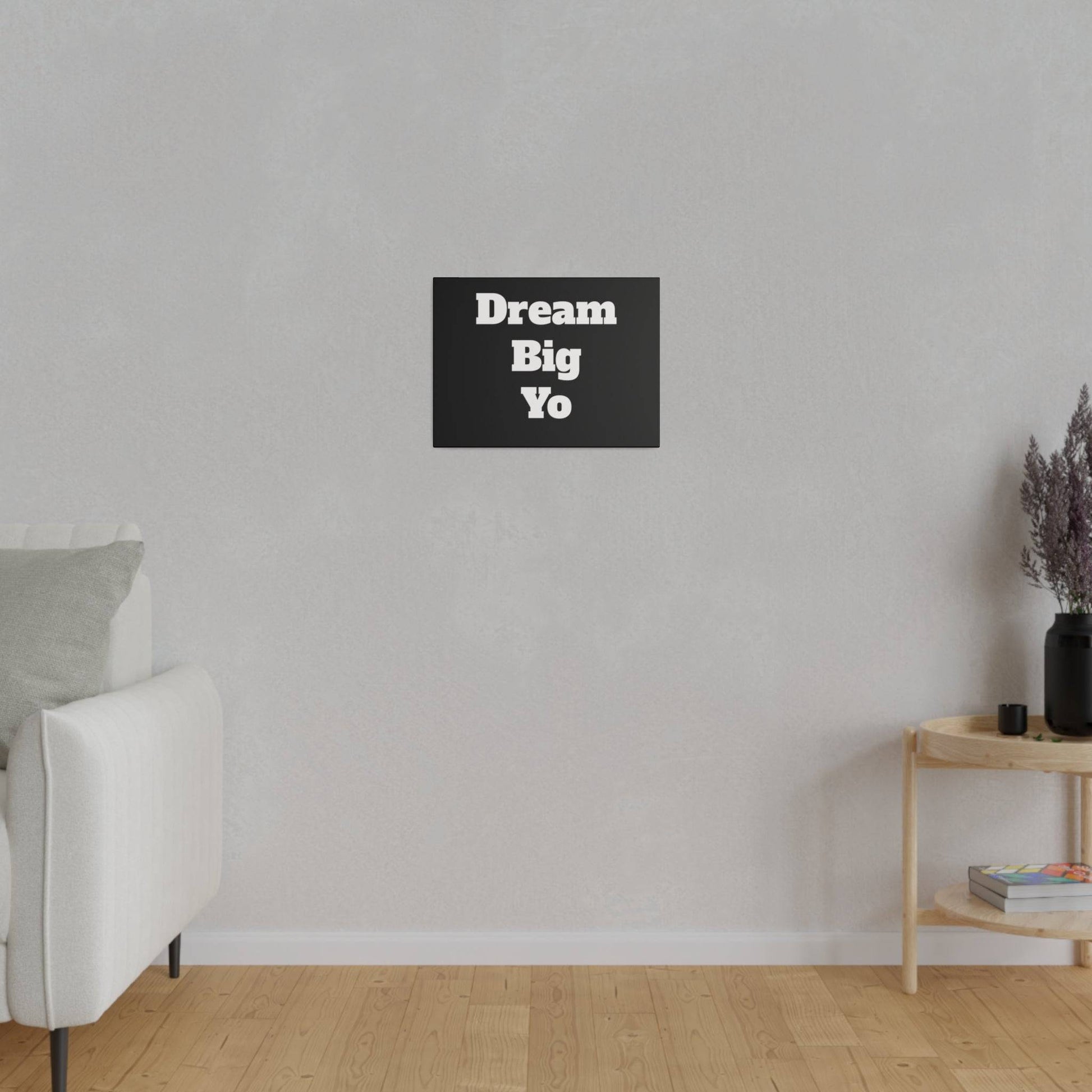 A minimalist living room with a light gray wall, white sofa on the left, and a black wall art sign reading “Dream Big Yo” centered above.