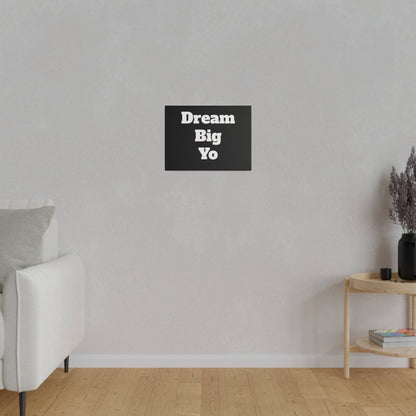 A minimalist living room with a light gray wall, white sofa on the left, and a black wall art sign reading “Dream Big Yo” centered above.