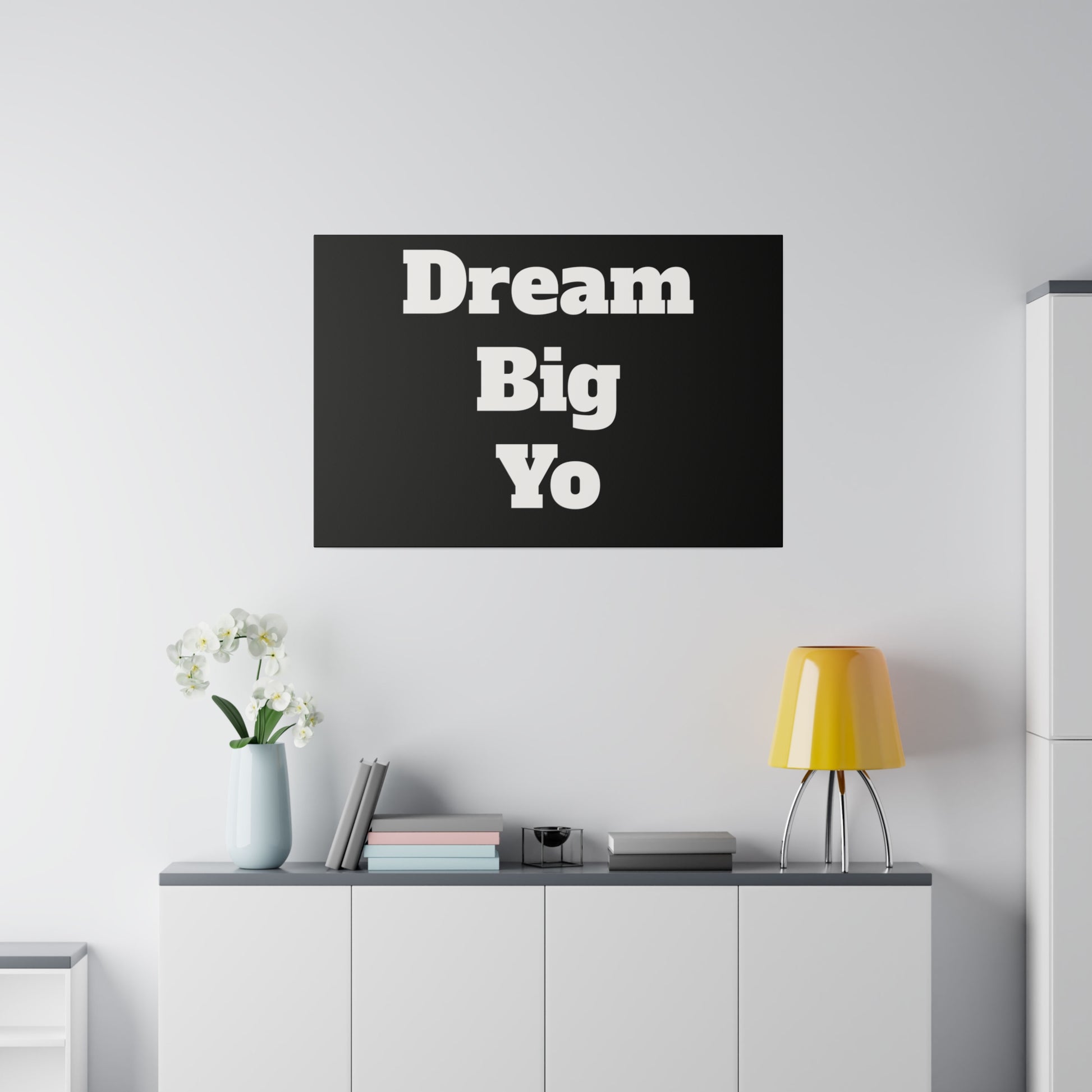 Black rectangular wall art with white stacked text 'Dream Big Yo' hangs above a white cabinet, with a yellow lamp and vase of white