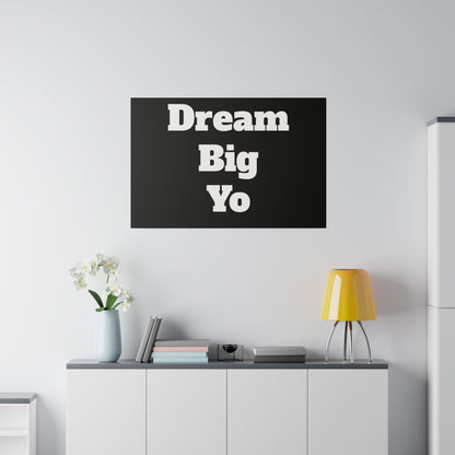 Black rectangular wall art with white stacked text 'Dream Big Yo' hangs above a white cabinet, with a yellow lamp and vase of white