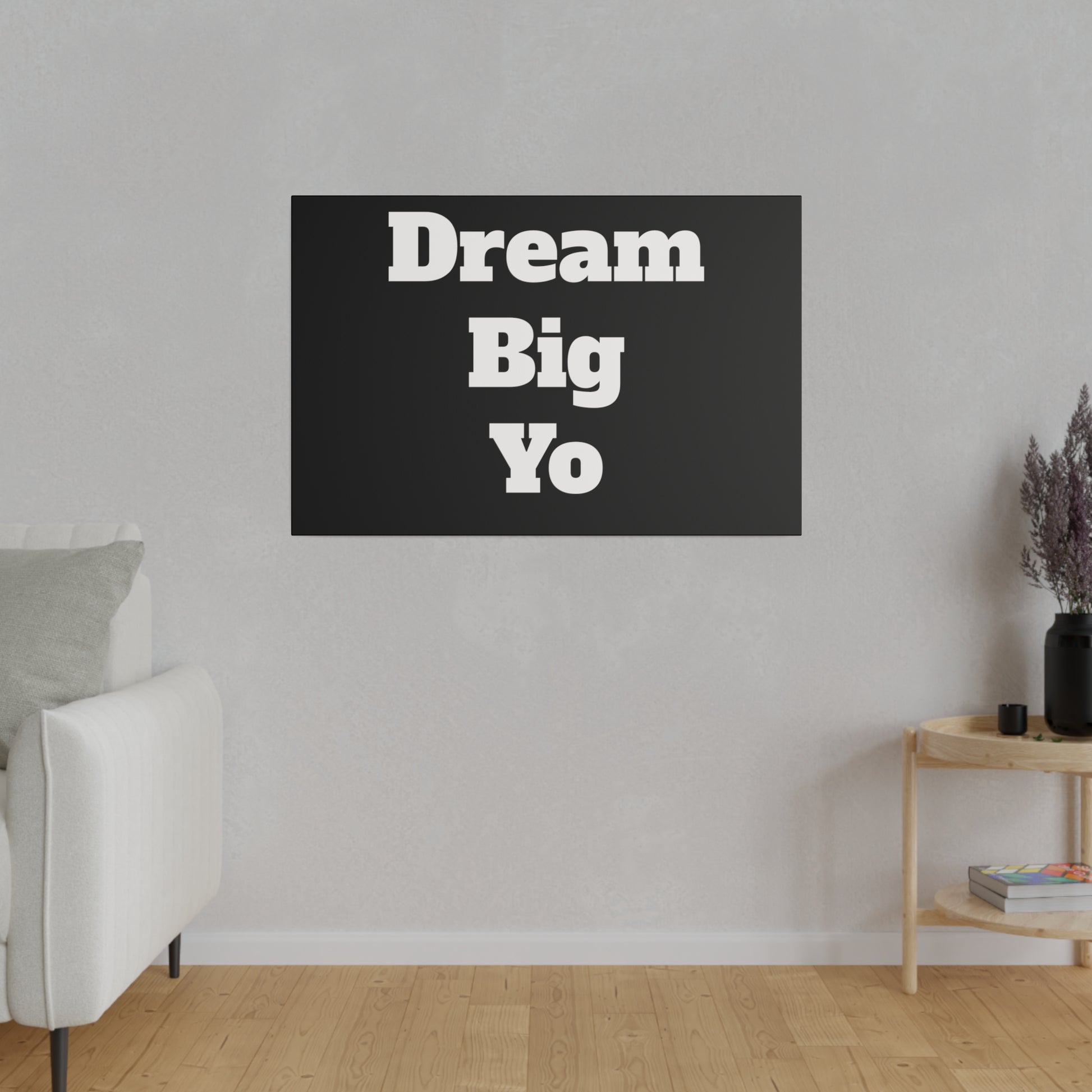 Black rectangular wall art with bold white text reading “Dream Big Yo” hanging above a light living room with a white sofa.