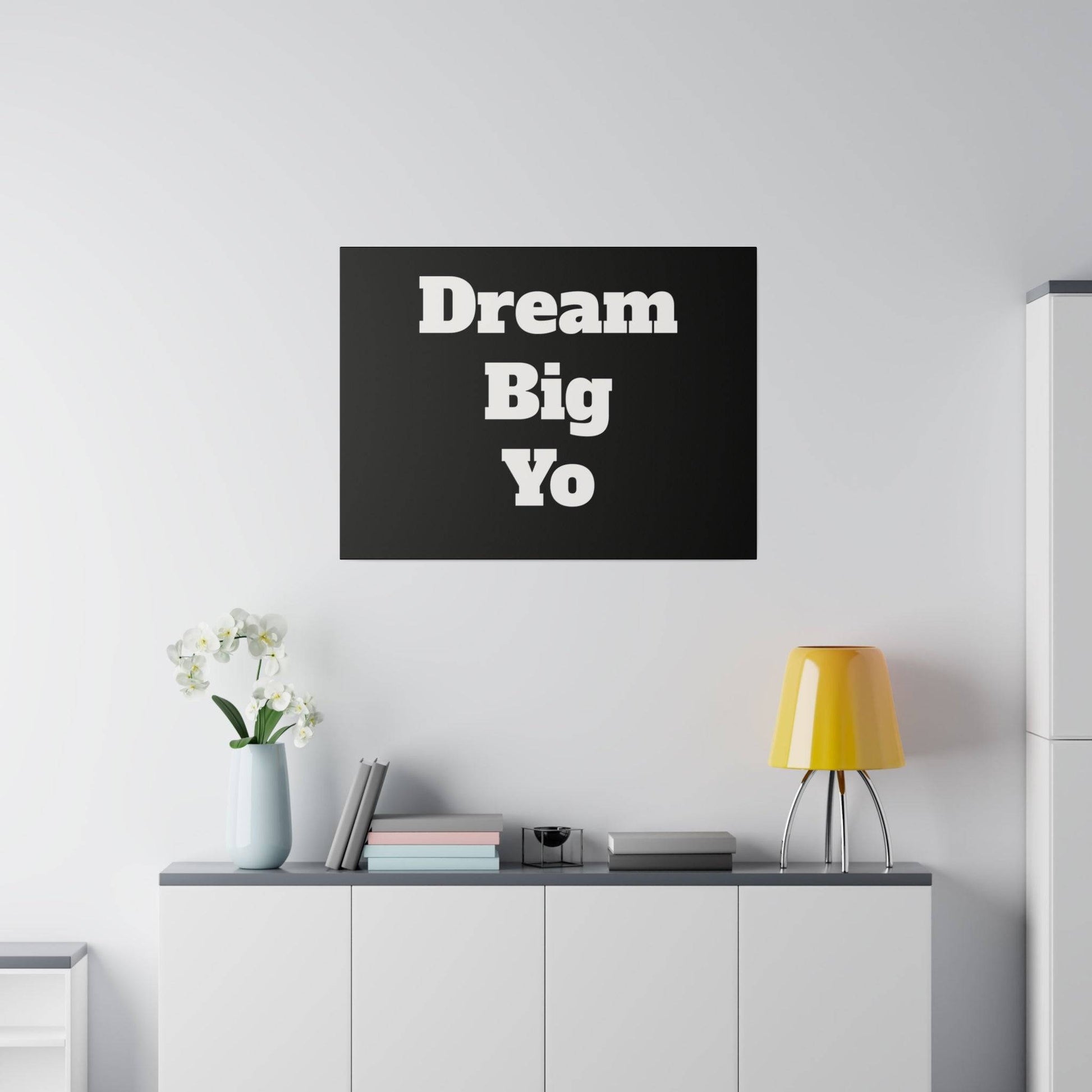 Black wall art with bold white text reading “Dream Big Yo” hangs above a white cabinet with a vase, books, and a yellow lamp.
