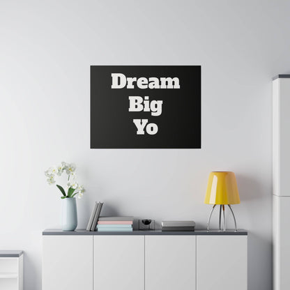Black wall art with bold white text reading “Dream Big Yo” hangs above a white cabinet with a vase, books, and a yellow lamp.