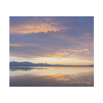 Calm lake at sunset with pastel orange and purple sky, snow-capped mountains on the horizon and their reflection on the water.