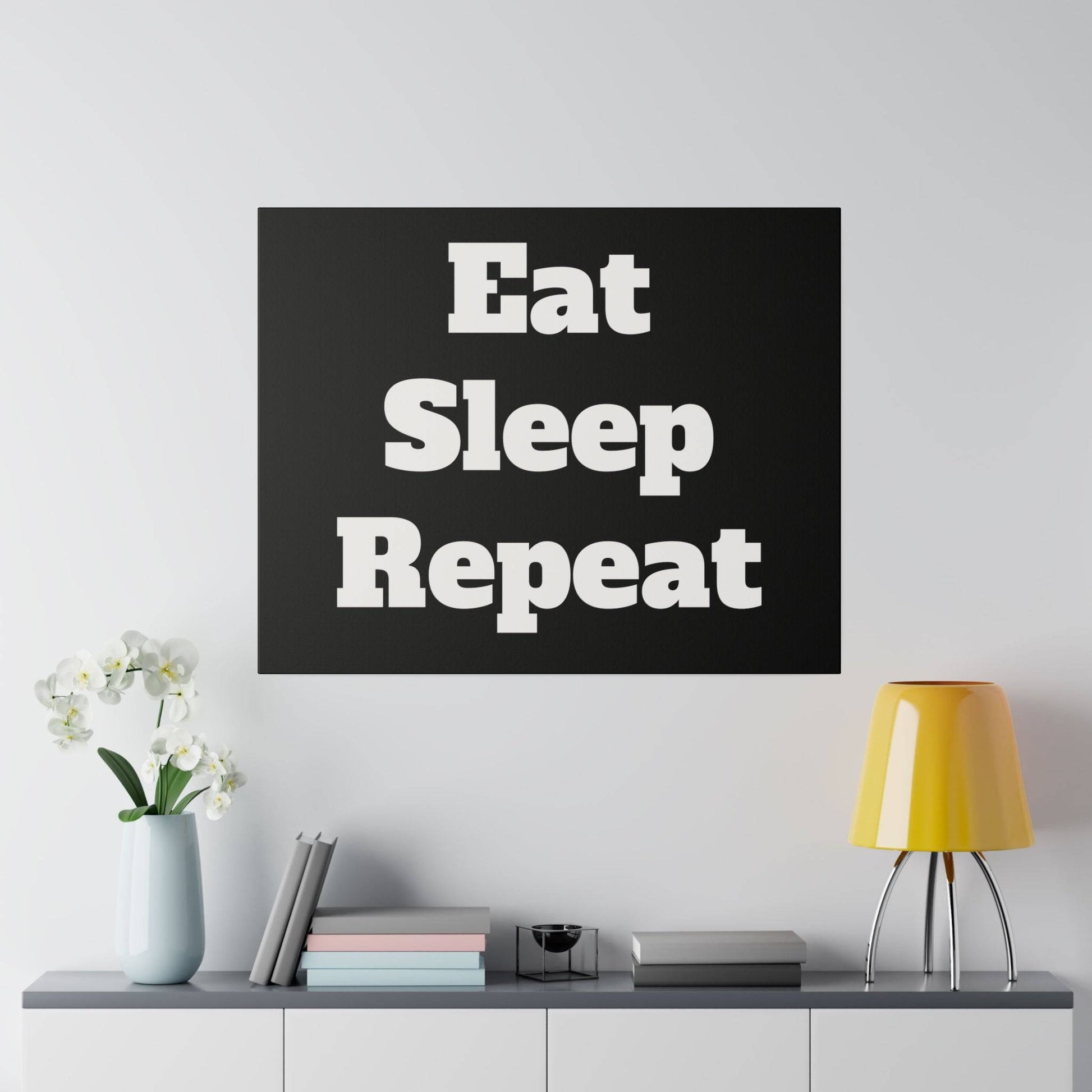 Black square wall art reading 'Eat Sleep Repeat' in white bold letters, hung above a gray cabinet with a yellow lamp and vase.