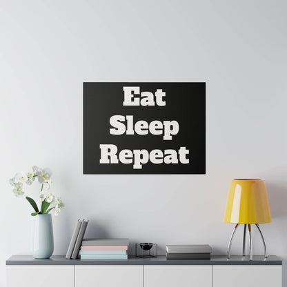 Black square wall art with white bold text reading “Eat Sleep Repeat,” hung above a console table with vase, books, and a yellow lamp.