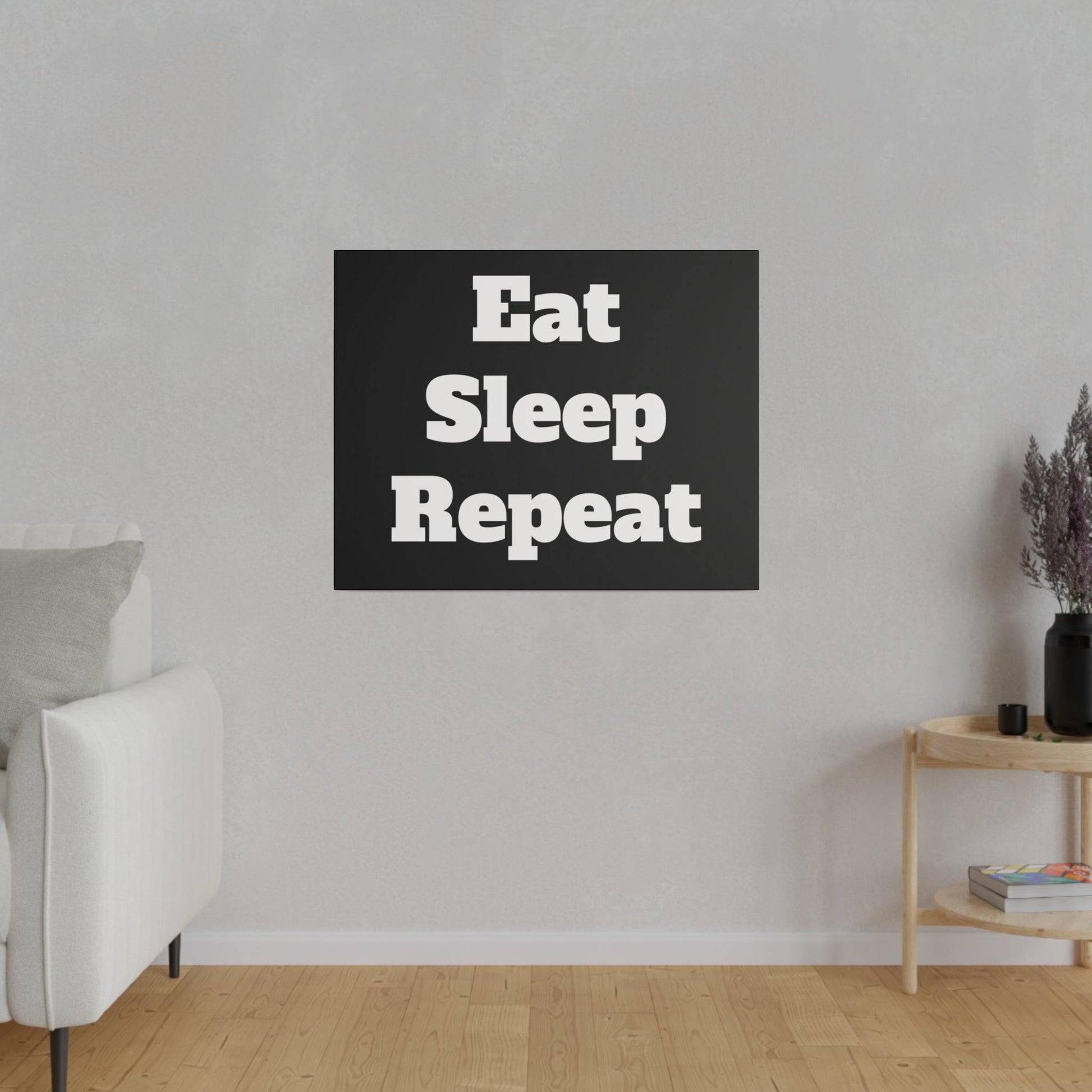 Black square wall art reading “Eat Sleep Repeat” in a modern living room with a beige sofa and a wooden side table.