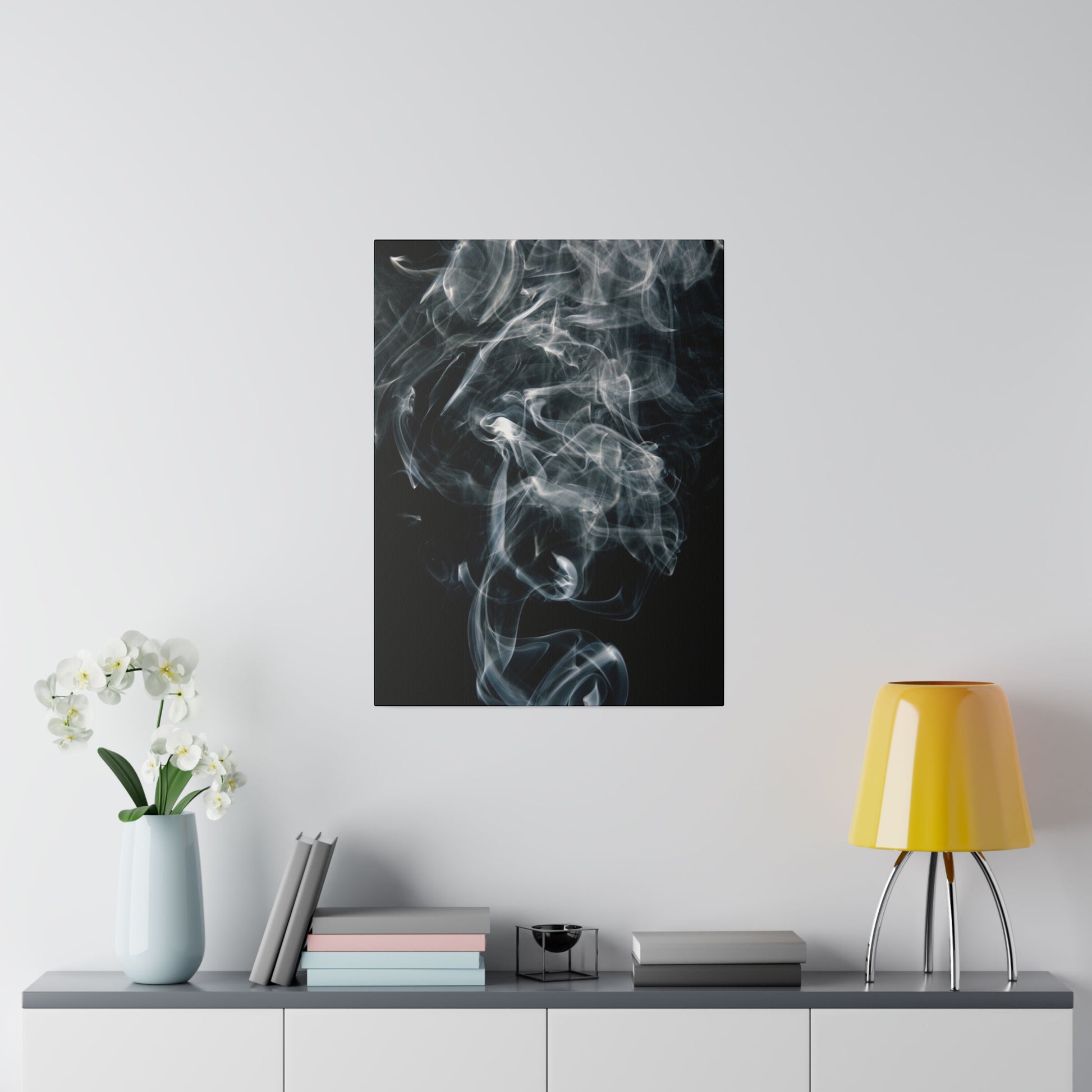 Vertical art print with white smoke on a black background, hung over a gray sideboard beside a yellow lamp and a vase of white orchids.
