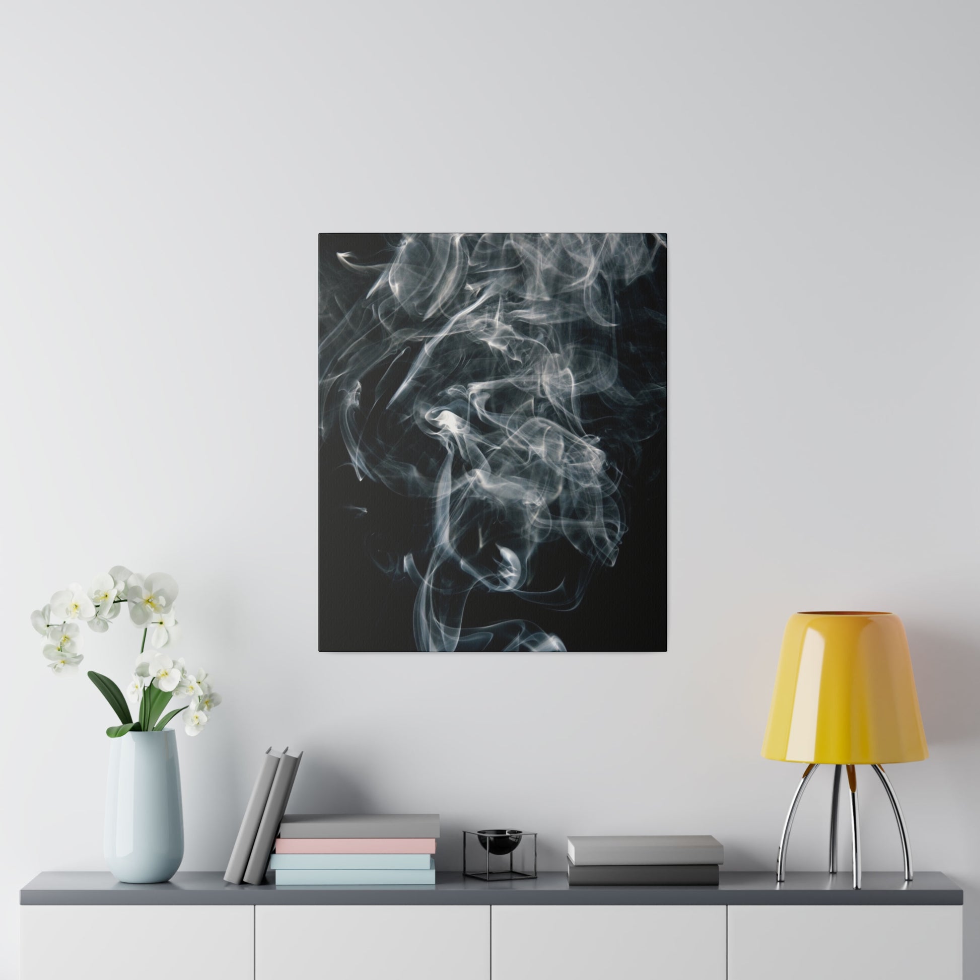 Square black canvas with wispy white smoke patterns hangs on a white wall above a gray sideboard with a light blue vase of white flowers