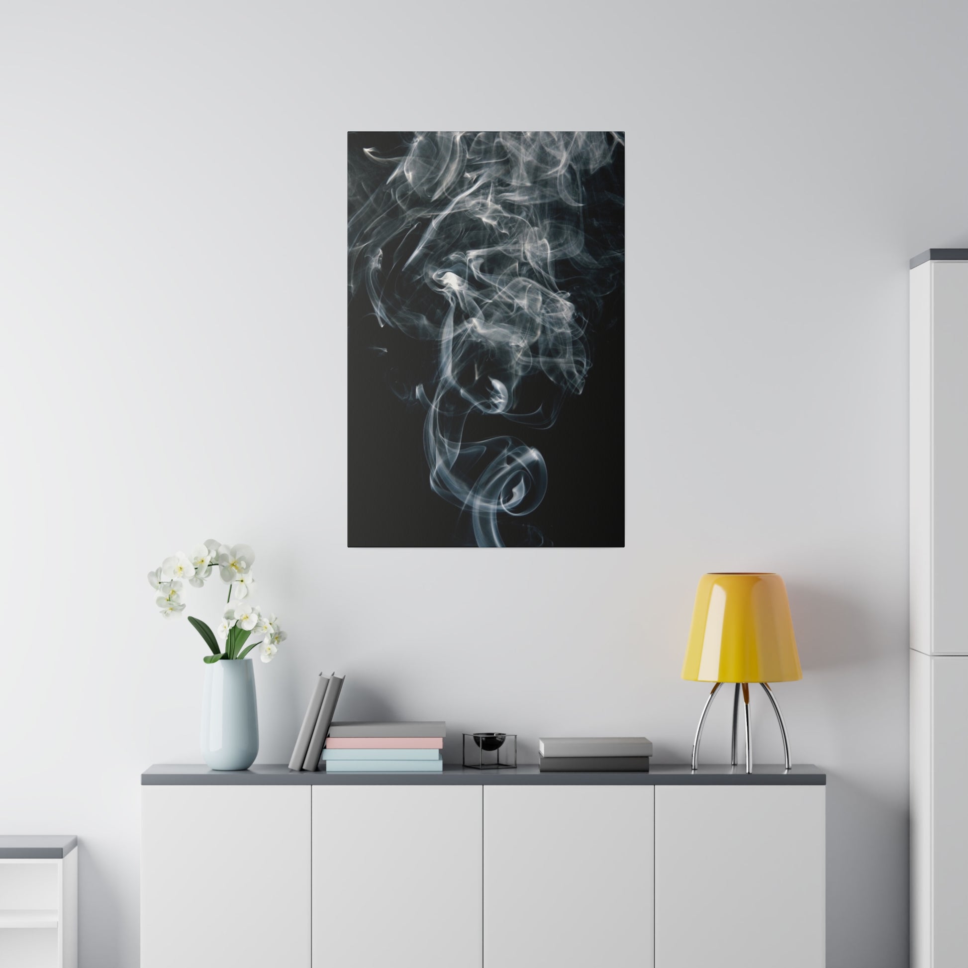 Black poster with white smoke pattern hanging above a white sideboard with a vase of orchids, books, and a yellow lamp.