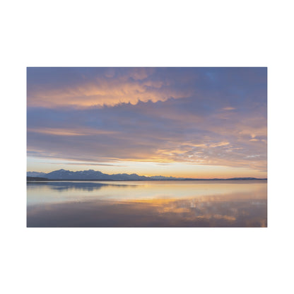 Calm lake at sunset with snow-capped mountains on the horizon and a colorful, cloud-filled sky reflected on the water.