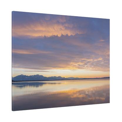 Wide lake at sunset reflects orange, pink, and blue sky with distant snow-capped mountains on the horizon.