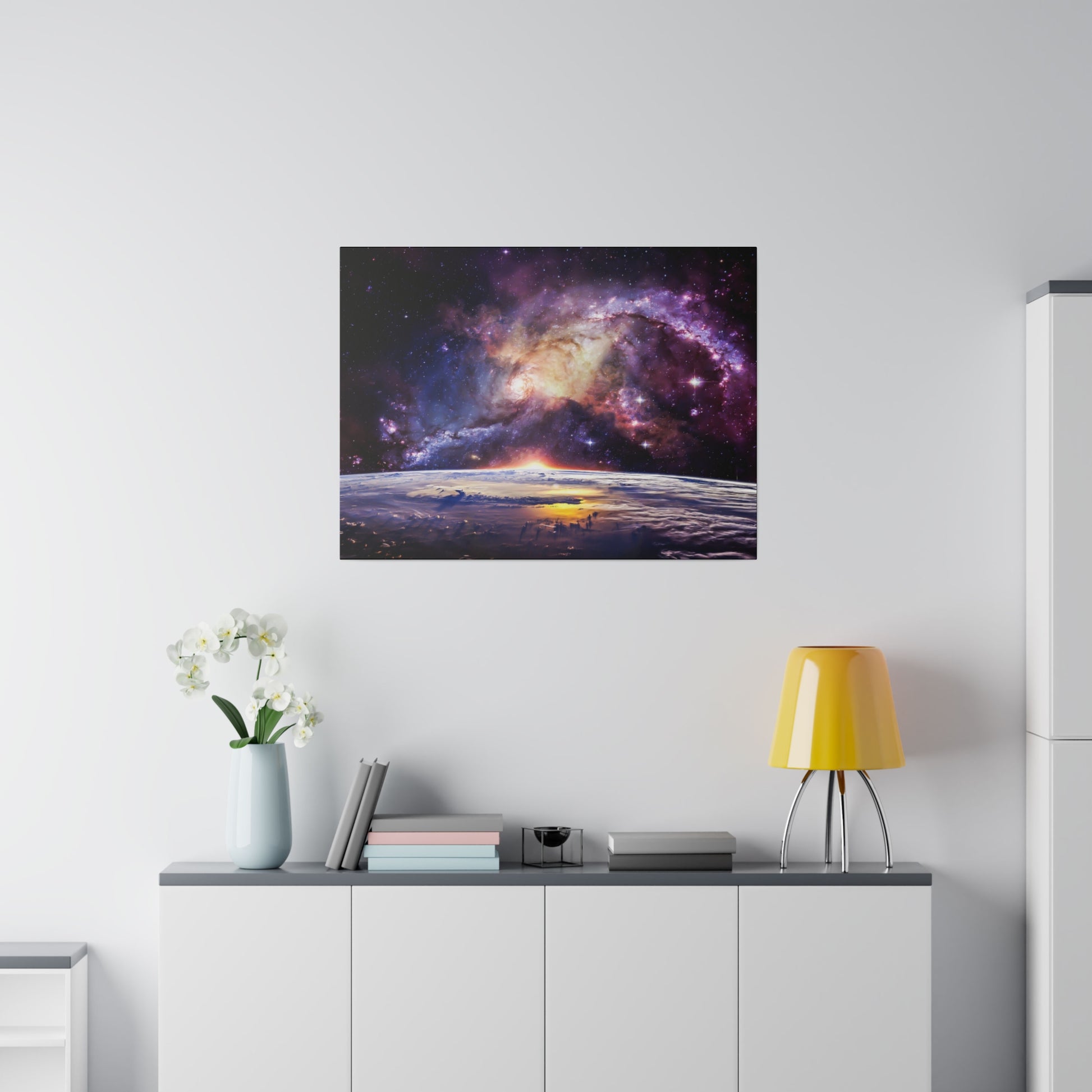 Square art print of a colorful galaxy above a white cabinet with a yellow lamp, books, and a vase of white flowers.