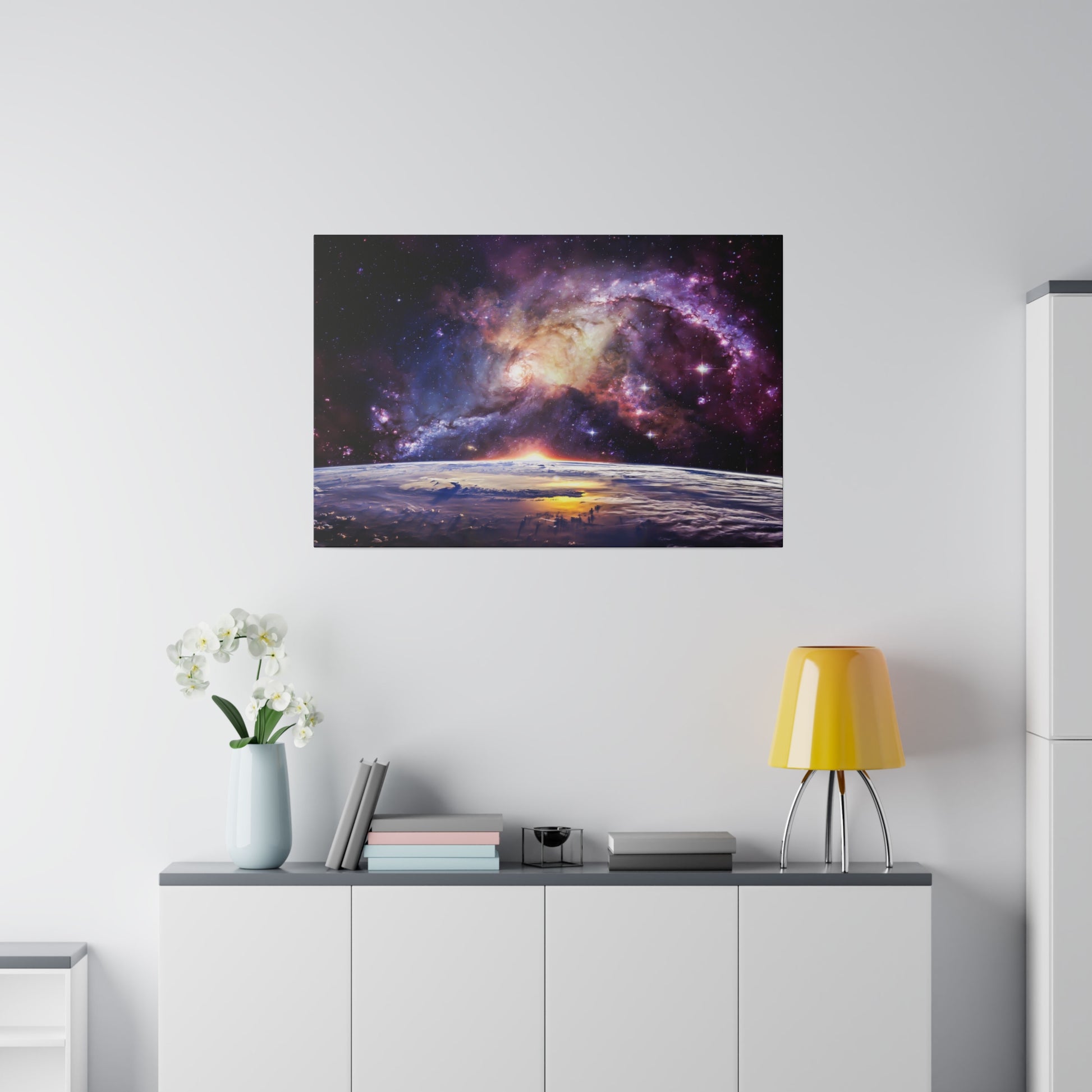 Space-themed artwork showing a colorful nebula over Earth, hung above a white cabinet with a yellow lamp and a vase of white flowers.