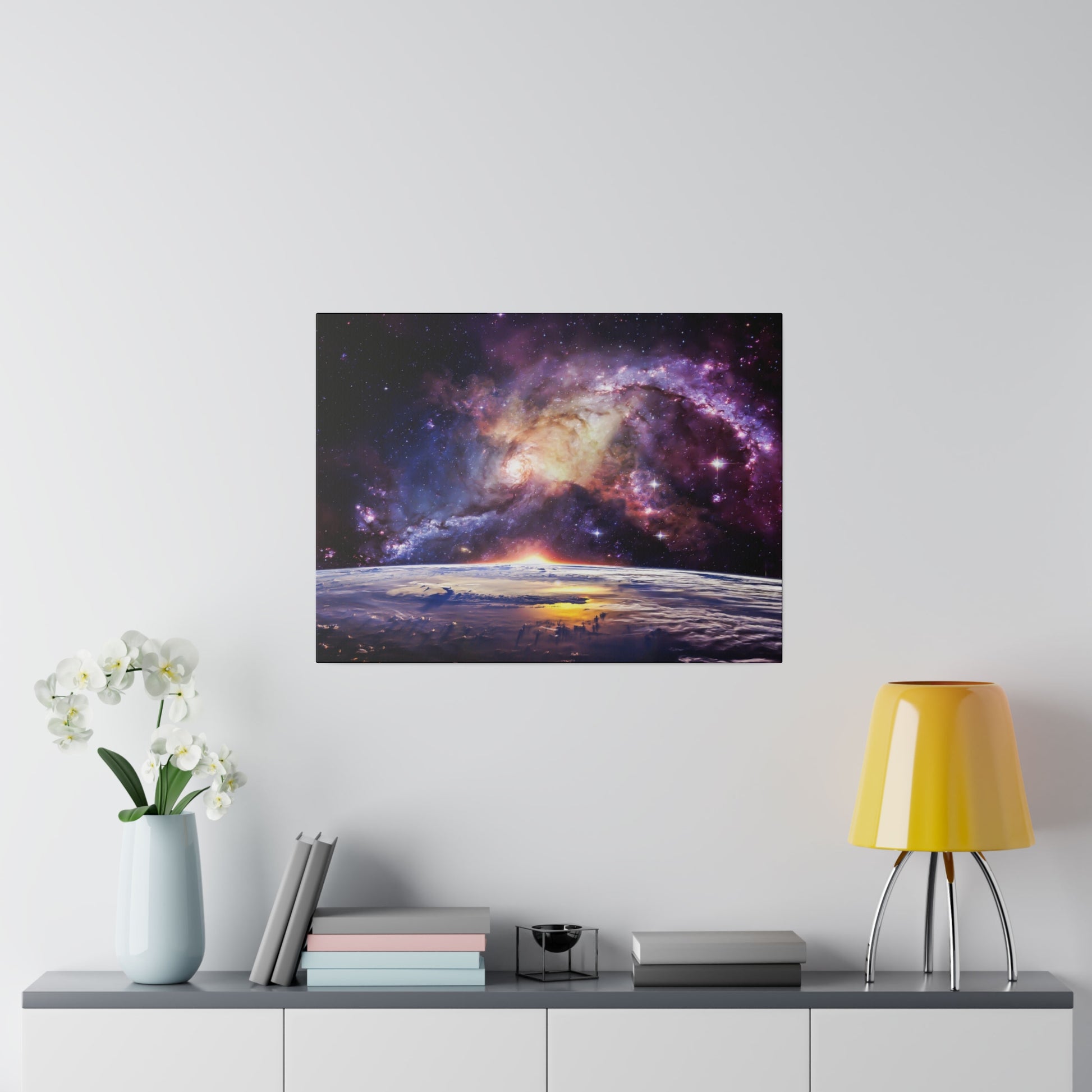 Square wall art showing a galaxy and sunrise over Earth’s horizon, mounted above a console with a yellow lamp and white flowers.