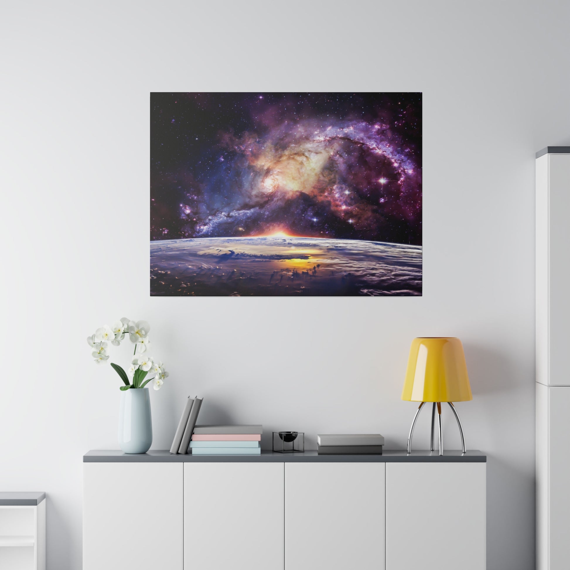 Square wall art of a colorful nebula above Earth’s horizon, mounted above a white cabinet with a yellow lamp and vase.