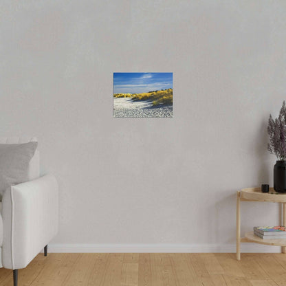 Small landscape painting of sandy dunes with yellow grasses under a blue sky, hung on a light gray wall.