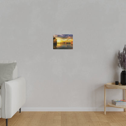 Small rectangular landscape painting of a sunset over a river, hung on a light gray wall above a wooden floor.