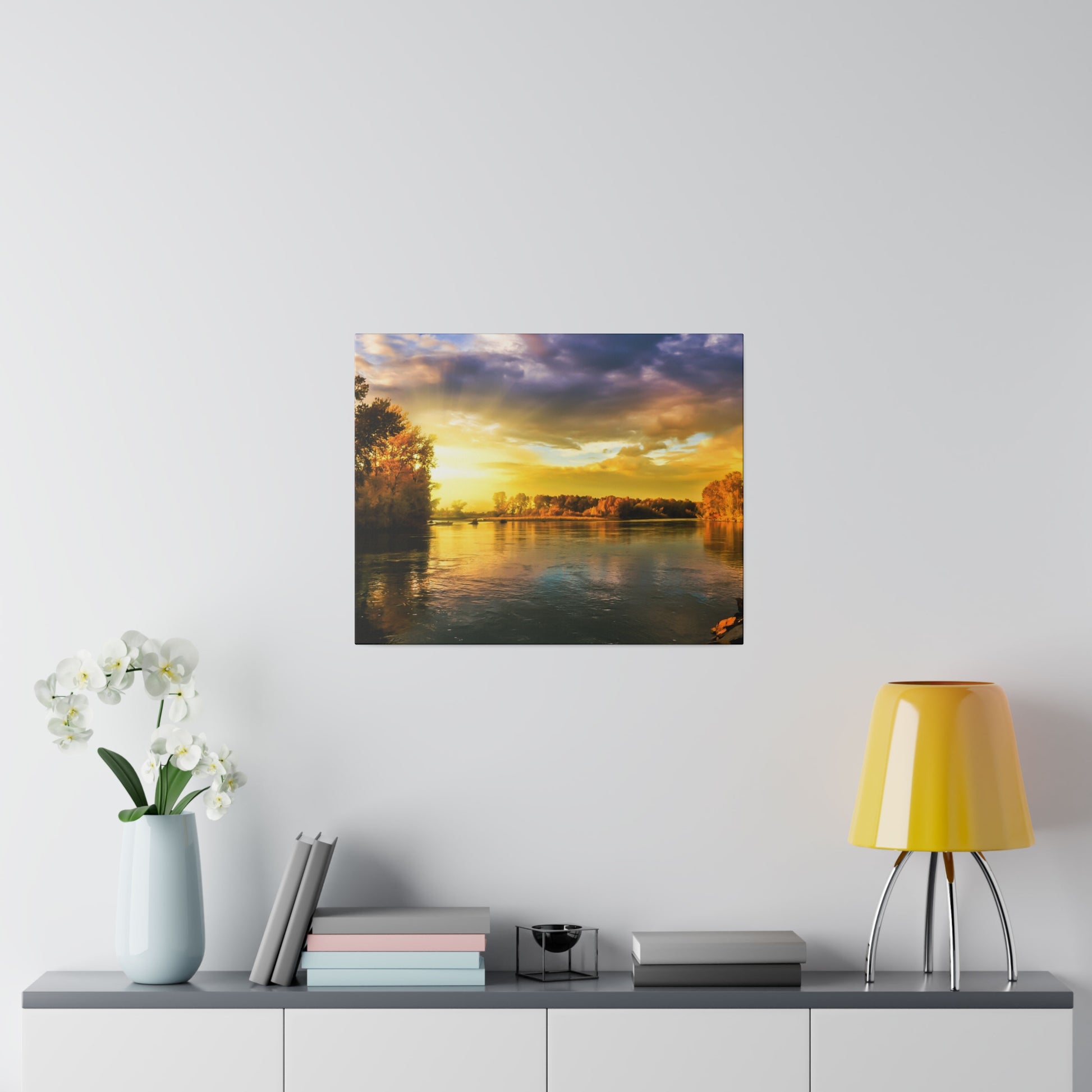 Square landscape print of a sunset over a river, mounted on a light wall above a cabinet with a vase of white orchids and a yellow lamp.