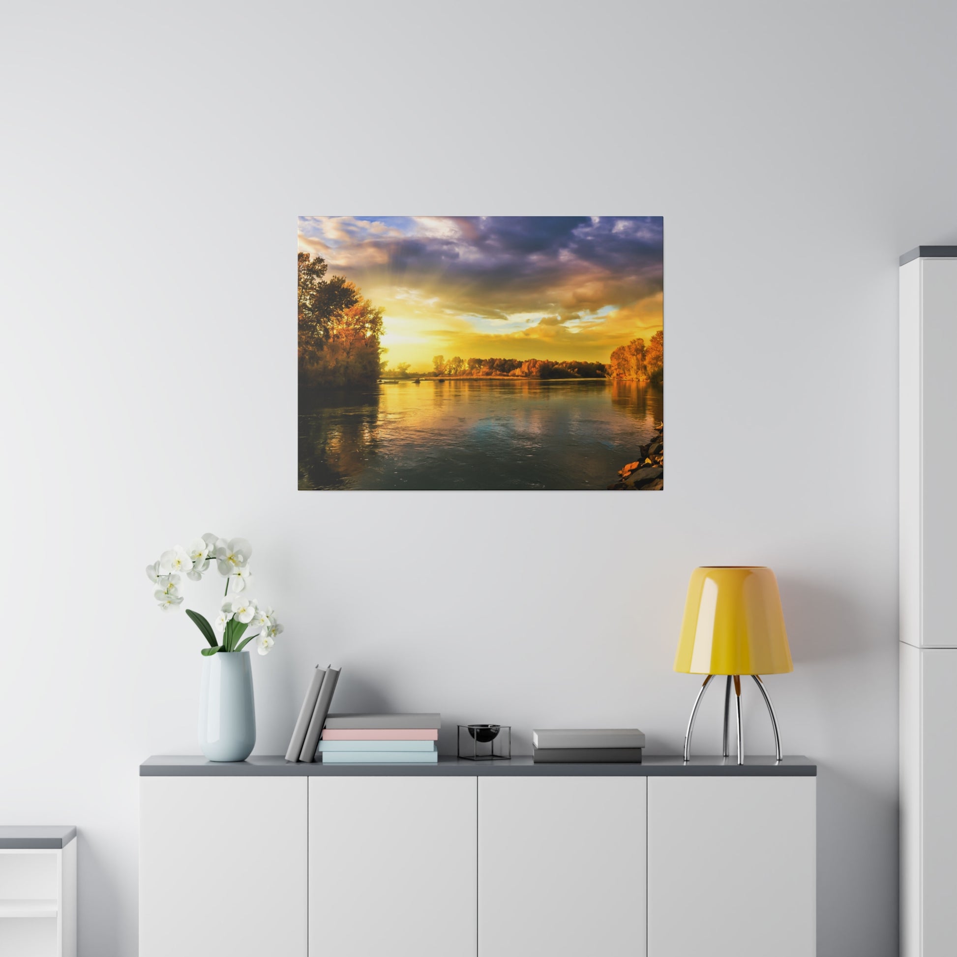 Square wall art showing a sunset over a calm river with autumn trees, hung above a white cabinet.