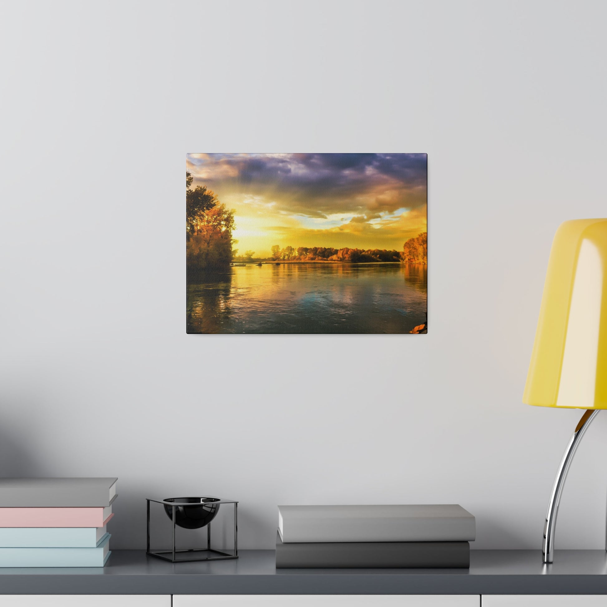 Framed sunset river landscape print on a light gray wall above a shelf with stacked books and a yellow lamp.