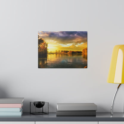 Framed sunset river landscape print on a light gray wall above a shelf with stacked books and a yellow lamp.