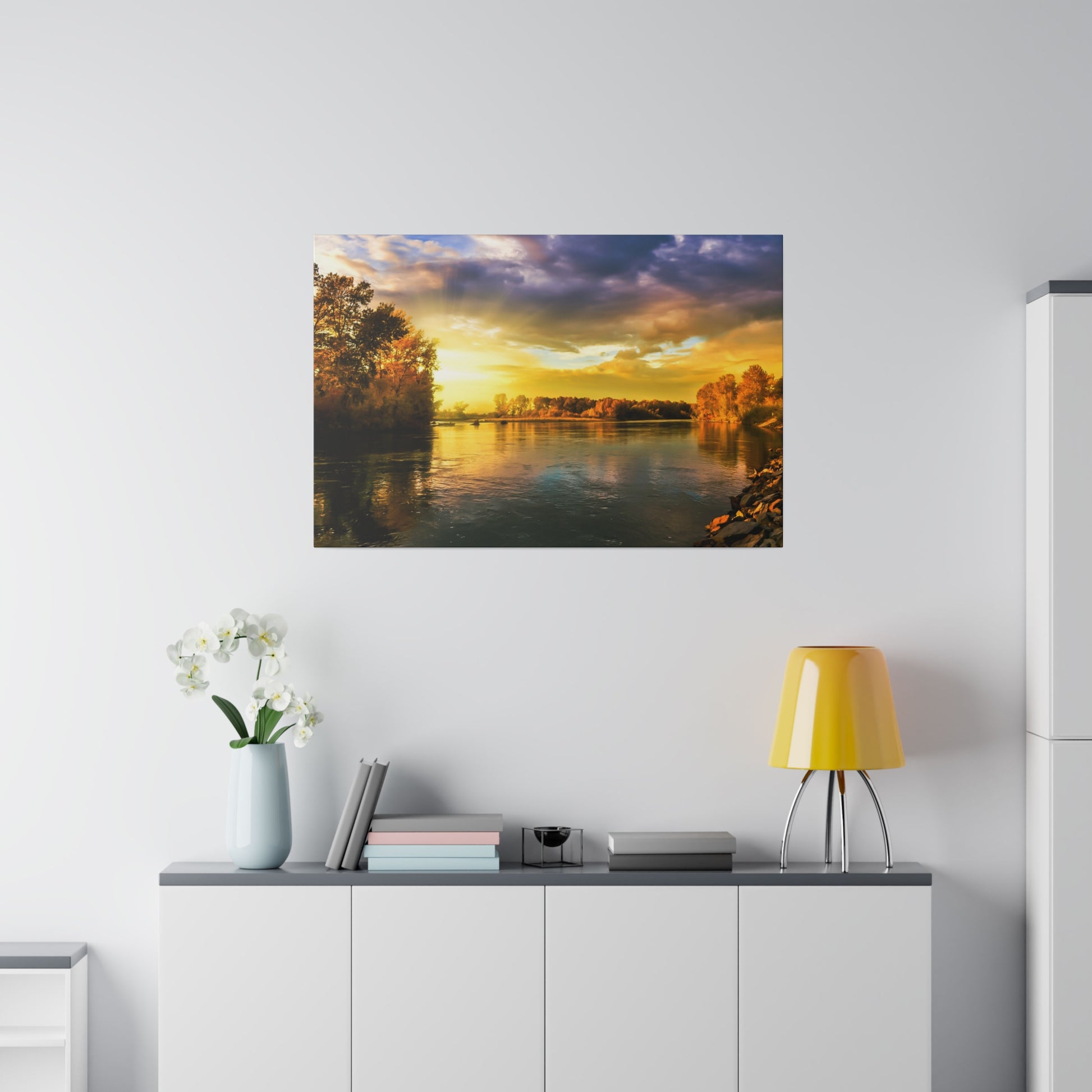 Framed sunset river landscape photograph on a white wall above a cabinet with a yellow lamp and white vase flowers.