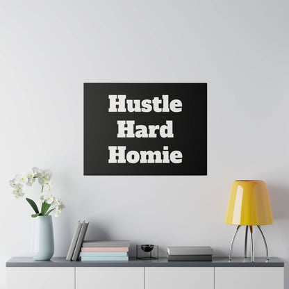 Black square wall art with bold white words “Hustle Hard Homie” hanging above a shelf with a vase and yellow lamp.
