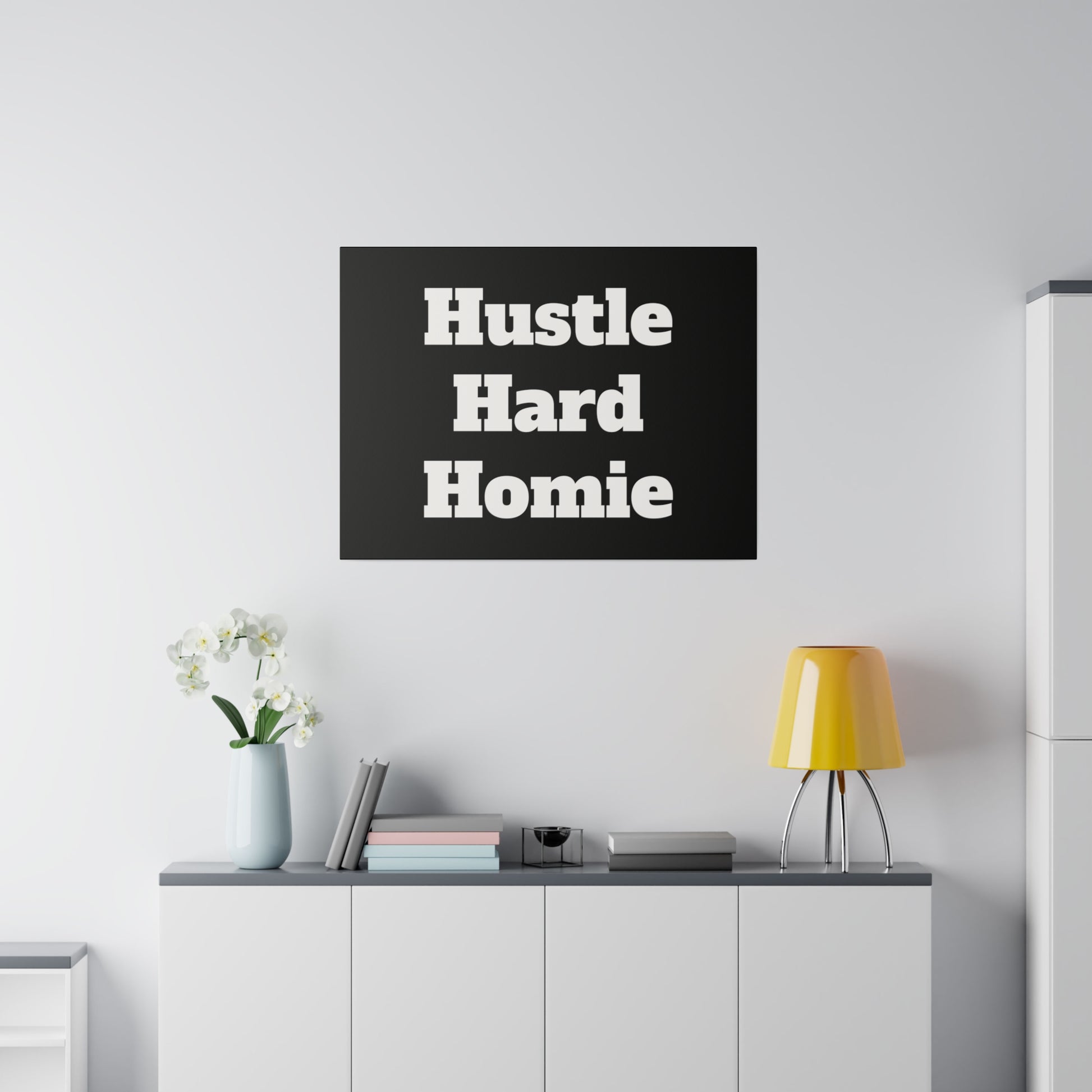 Black square poster with bold white text “Hustle Hard Homie” on a wall above a gray cabinet with books, vase, and a yellow lamp.