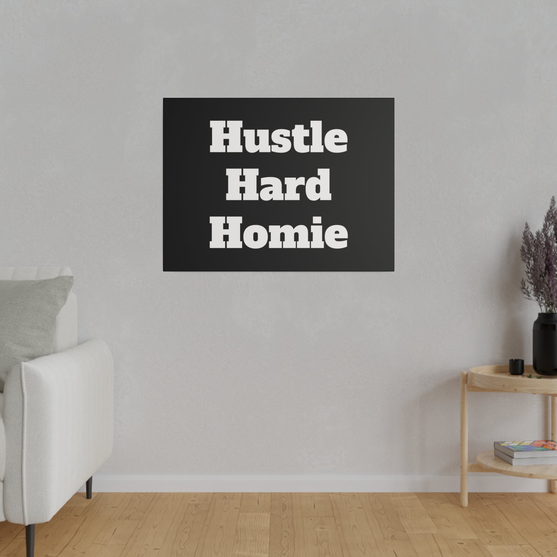 Black rectangular wall art with bold white text "Hustle Hard Homie" hung on a light gray living room wall beside a white sofa and a wooden