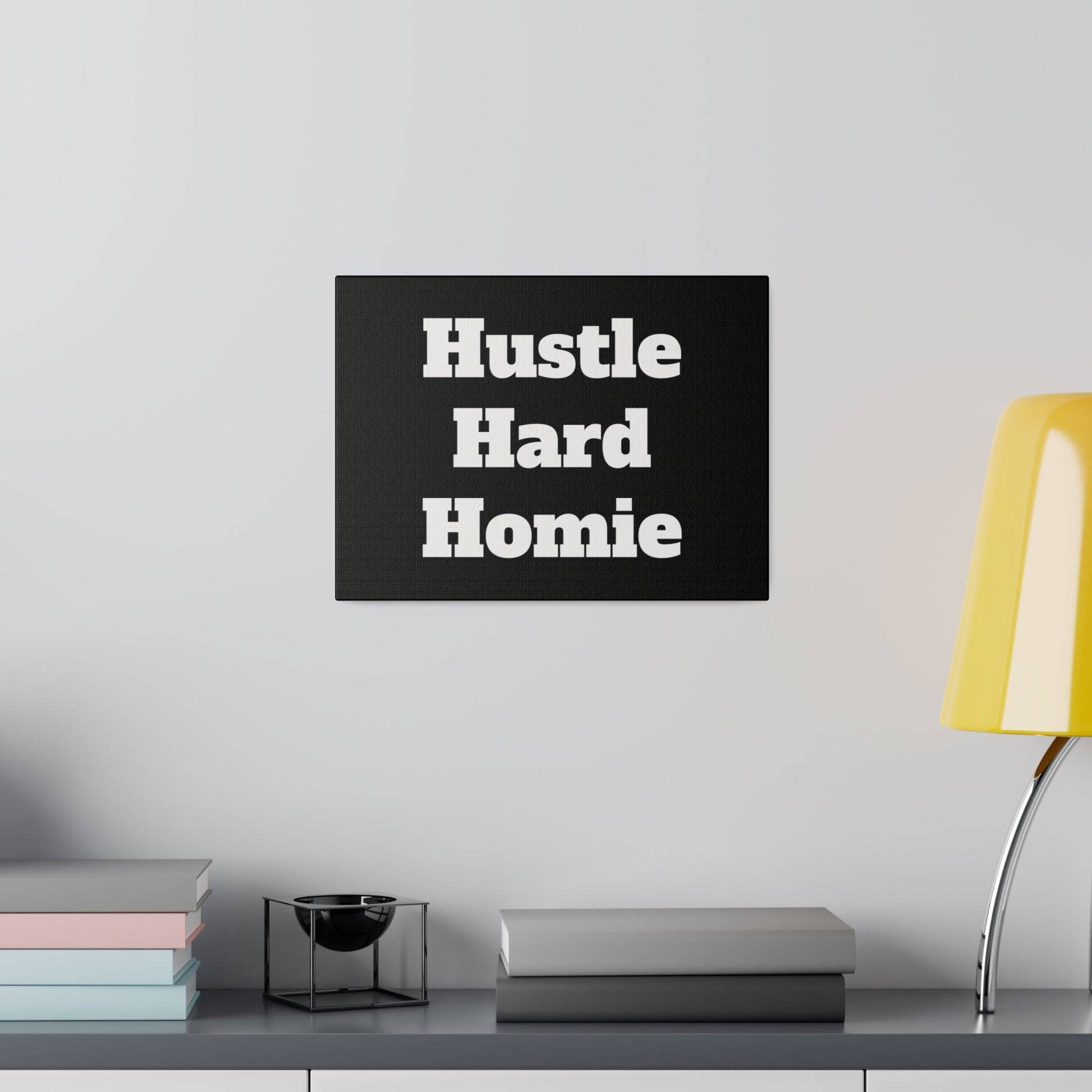 Black rectangular wall art with white bold text "Hustle Hard Homie" mounted over a shelf with pastel book stacks, a black decorative bowl,