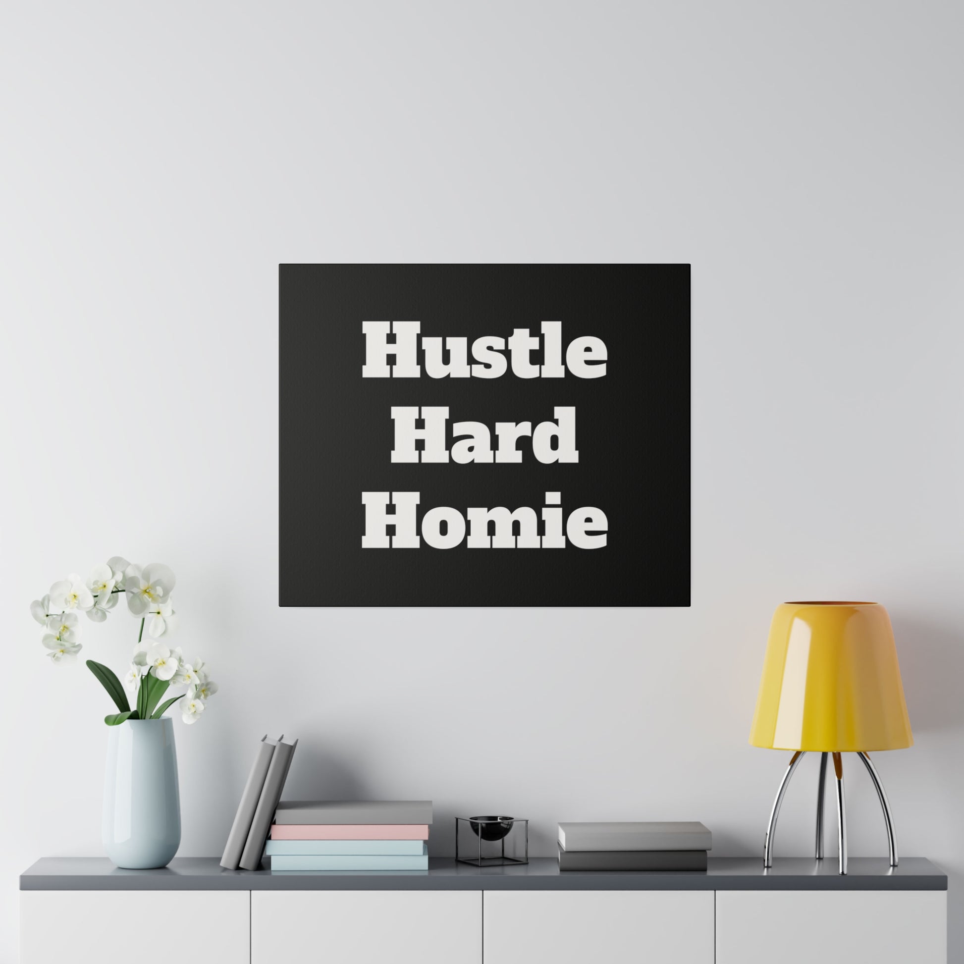 A black square wall art with the phrase “Hustle Hard Homie” in large white letters above a gray sideboard with a light blue vase of white