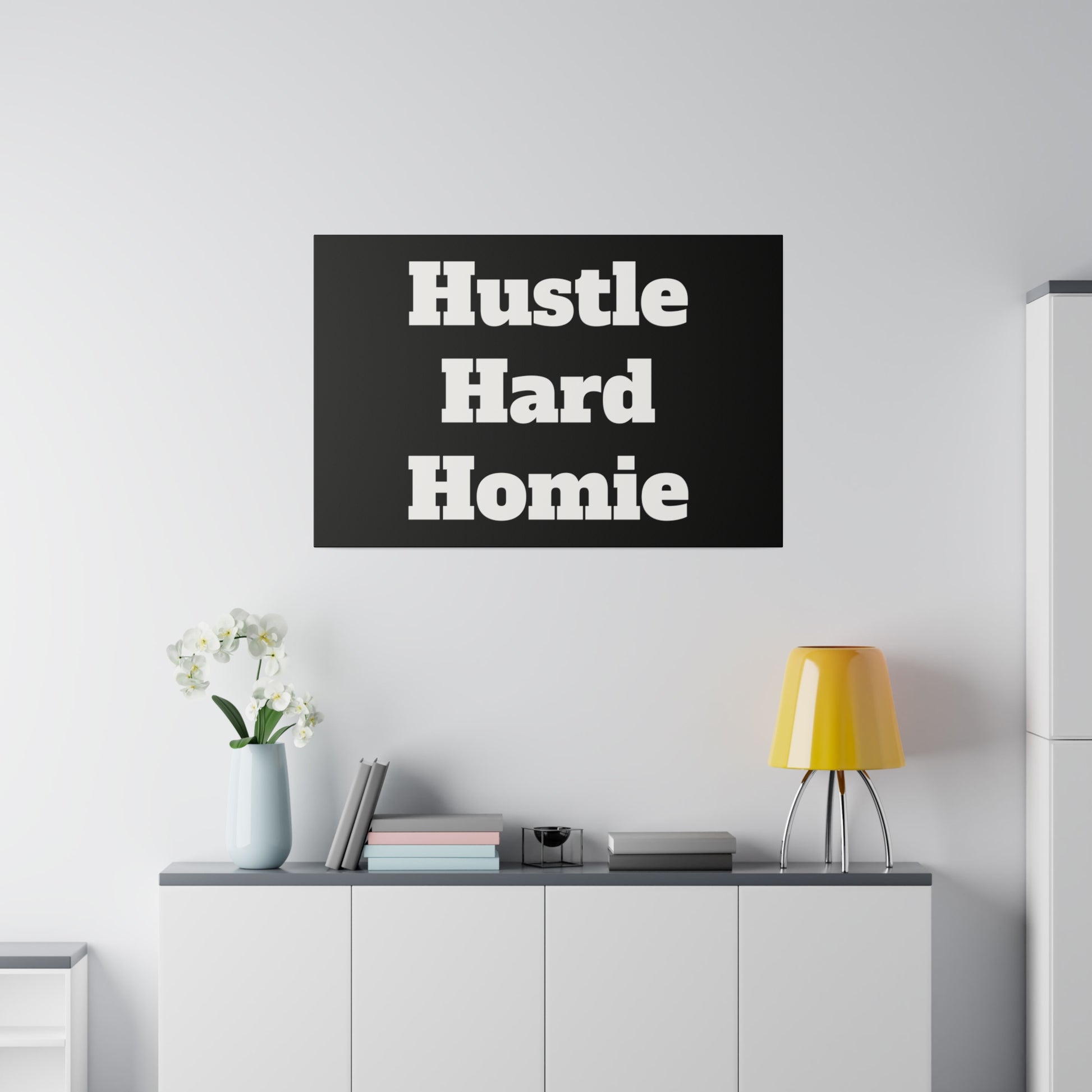 Black rectangular poster with bold white text "Hustle Hard Homie" hung above a white cabinet with a yellow lamp.