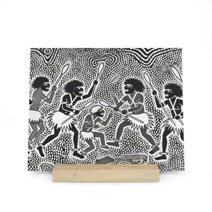 Square black-and-white artwork showing stylized tribal dancers with spears and grass skirts, mounted on a light wooden stand.