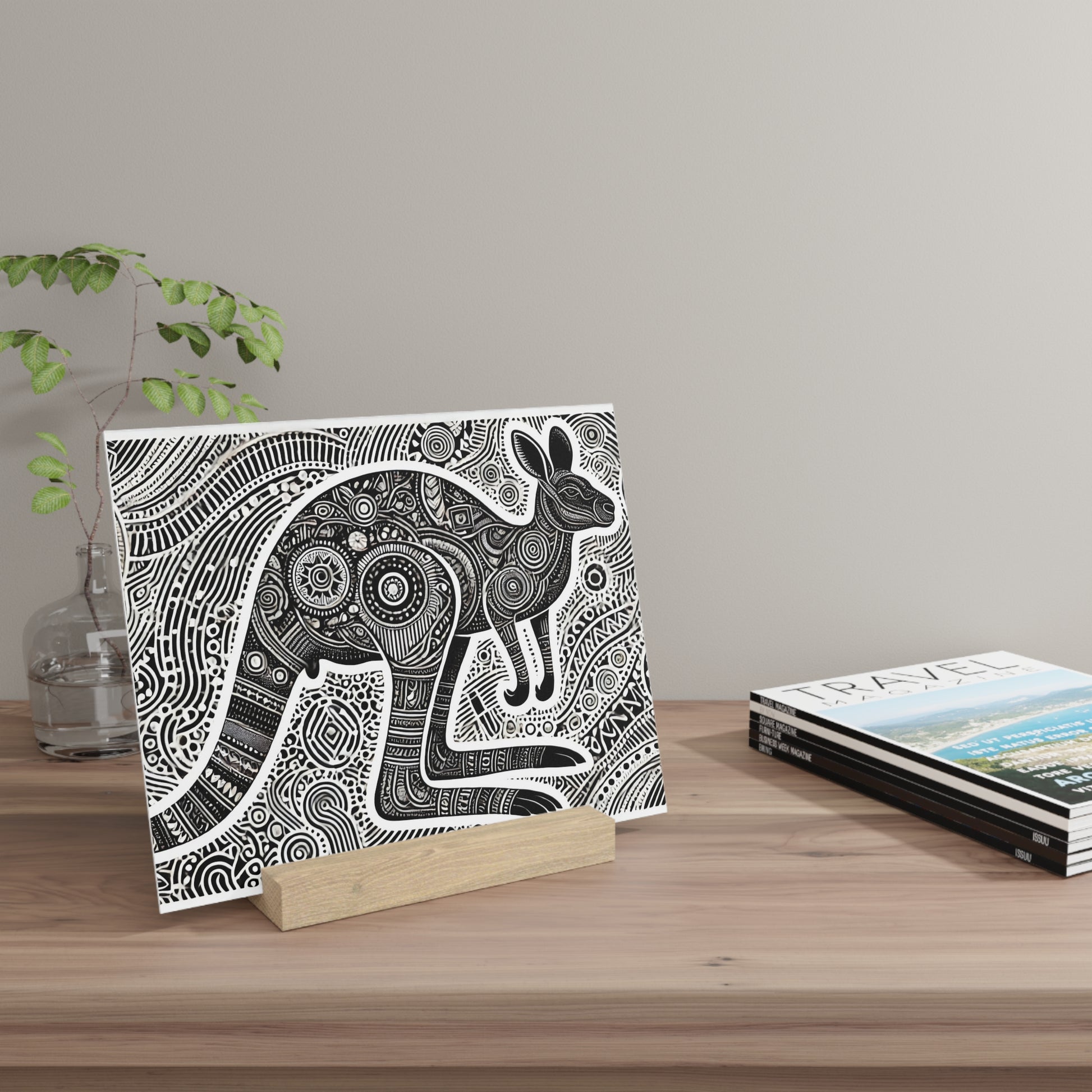 Black and white intricate elephant illustration on a wooden stand, placed on a light wood table beside a glass vase and travel magazines.