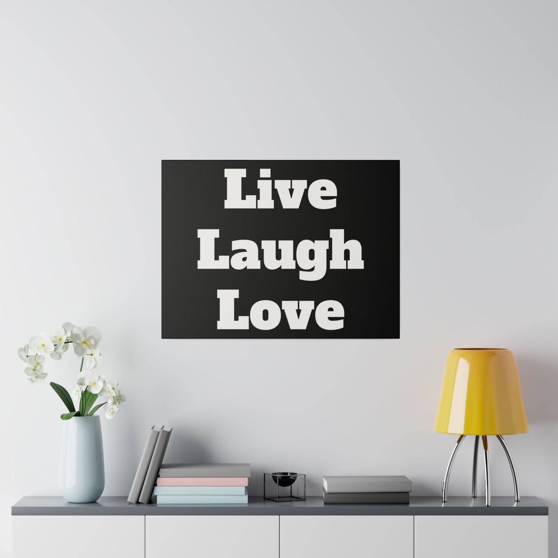 Black square wall art with white “Live Laugh Love” text hangs above a gray shelf with a vase, books, and a yellow lamp.