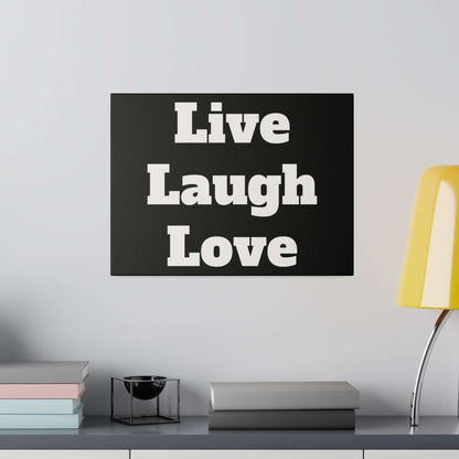Black rectangular canvas with white 'Live Laugh Love' typography, mounted on a light wall above a shelf with books and a yellow lamp.