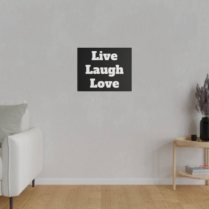 Black square wall art with bold white text reading “Live Laugh Love,” mounted on a light gray wall above a wooden floor.