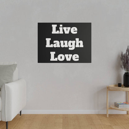 Black rectangular wall art reading 'Live Laugh Love' in white bold letters, hung on a light gray wall above a wooden floor in a living room.