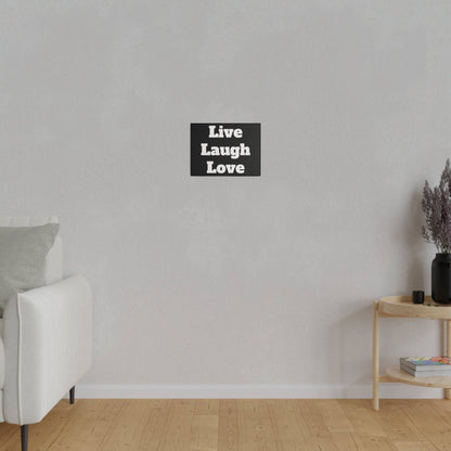 Small black square wall sign reading "Live Laugh Love" on a pale gray wall above a light gray sofa and wooden side table.